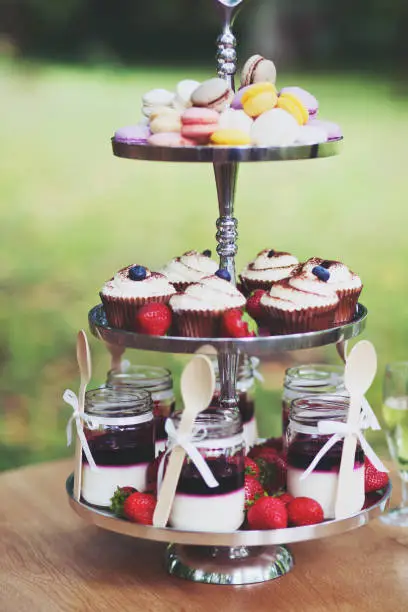 Desserts on a tiered platter - cupcakes with icing, panacota cream in jars, colourful macaroons - close up view.
