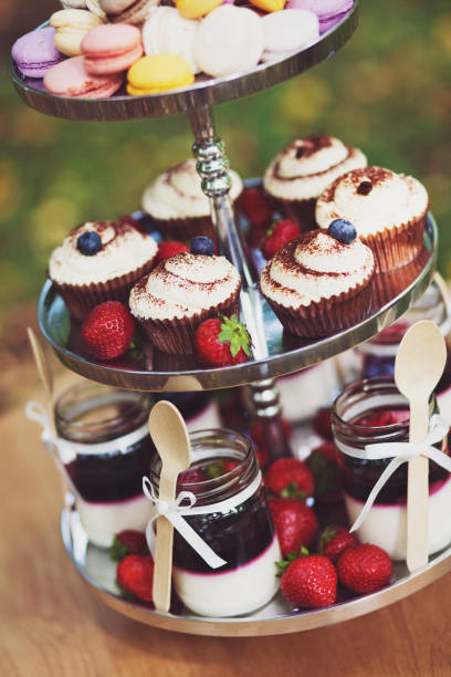 Close up of colourful vibrant three-tier tray full of cream cupcakes, macaroons, jars with yogurt and jam, strawberries - afternoon tea dessert platter. stock photo