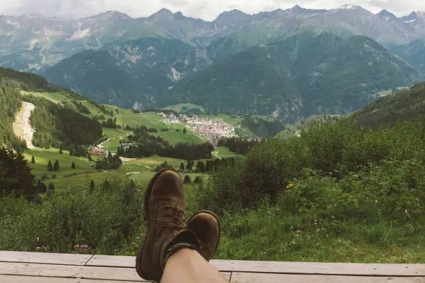 July 19, 2020 - Fiss-Ladis, Austria: rugged and worn hiking boots of a lying man ober the landscape and panorama of the mountains and valley and village below - Fiss_ladis, Austria, summer.