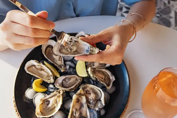 Woman eating fresh oysters with lemon close-up. Table with big plate of oysters and pink sparkling wine in a glass