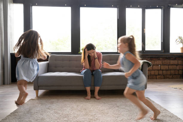 istock Young tired mother suffers from headache closed eyes touch forehead sitting on couch while her daughters running around 1271689506