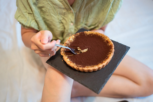 A top view of young pretty Asian woman eating delicious chocolate tart on bed with fork
