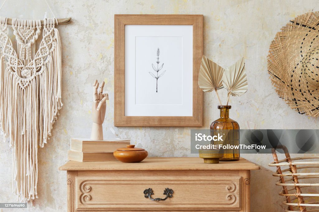 Stylish boho interior of living room with brown mock up poster frame, elegant accessories, flowers in vase, wooden shelf and hanging rattan hut. Minimalistic concept of home decor. Template. Beige boho interior of living room with mock up poster frame, elegant accessories in stylish home decor. Template. Wabi sabi concept. Home Decor Stock Photo