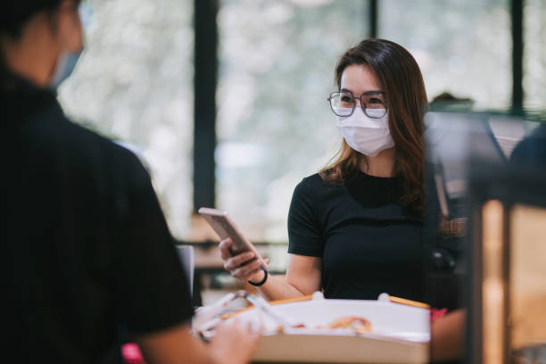 asian chinese beautiful woman making payment through contacless payment using her mobile app at the cashier with face mask in cafe