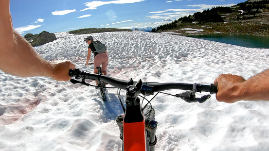 Young woman looks back and smiles to friend riding through snow, alpine lake below