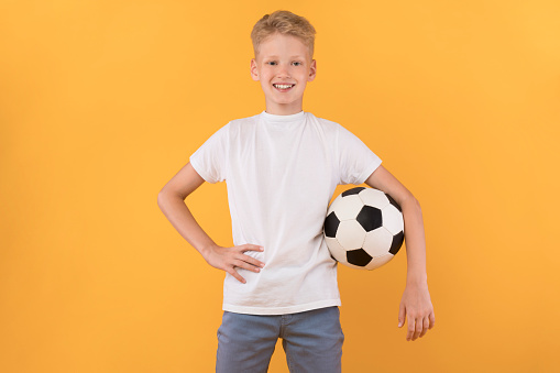 Football Player. Confident boy posing with soccer ball on pastel orange studio wall, banner, copy space