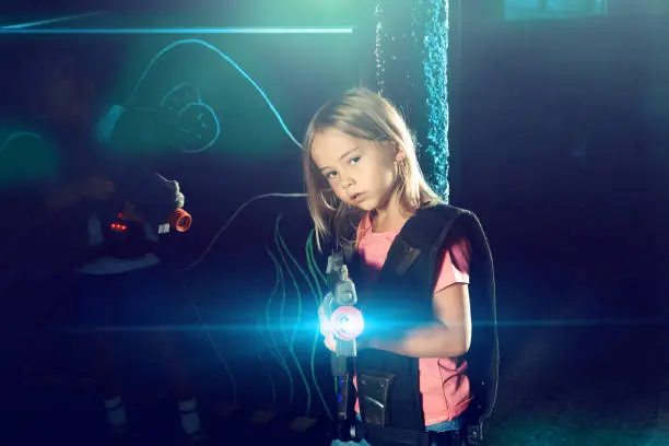 Positive little girl aiming laser gun at other players during lasertag game on dark arena