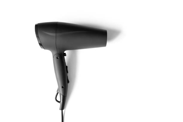 Young beautiful woman with long brown hair in a bathrobe is sitting on the bed and drying her hair with a hair dryer after bath. Morning routine. Ready for a new day.