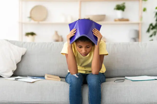 Upset Teen Girl Covering Head With School Exercise-Book Having Bad Grades Sitting On Sofa At Home. Teen Problems Concept