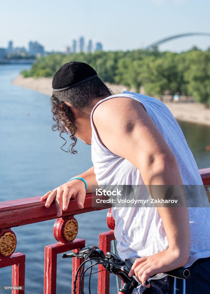 Unrecognizable young jewish man wearing twisted payot in black velvet kippah standing on the bridge and looking down into river. Vertical orientation. Unrecognizable young jewish man wearing twisted payot in black velvet kippah standing on the bridge and looking down into river. Vertical orientation Hasidism Stock Photo