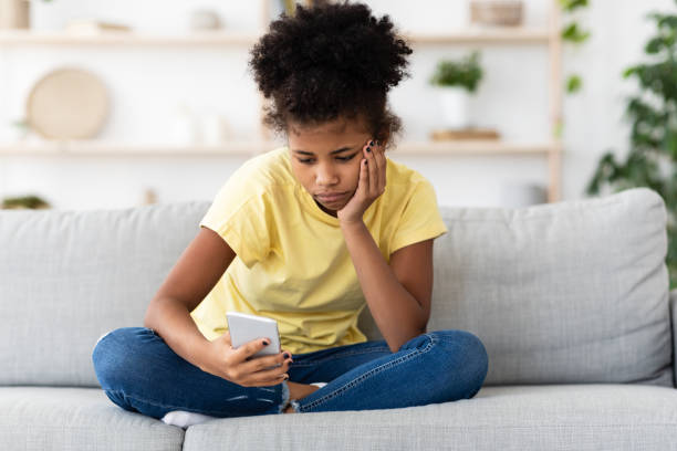 ragazza adolescente afroamericana annoiata che usa lo smartphone seduto a casa - cyberbullismo foto e immagini stock