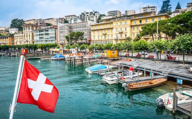 ルガーノ・ティチーノ湖の海岸にボートを持つスイスの国旗とルガーノの街並み - lugano ストックフォトと画像