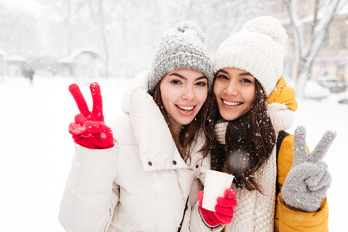 Portrait of two beautiful girls in warm wear smiling to camera and showing peace gesture in park in winter