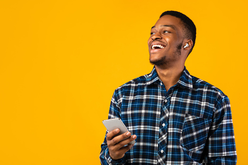 Happy African American Man Talking On Cellphone Hands-Free Using Earbuds Standing On Yellow Background. Studio Shot