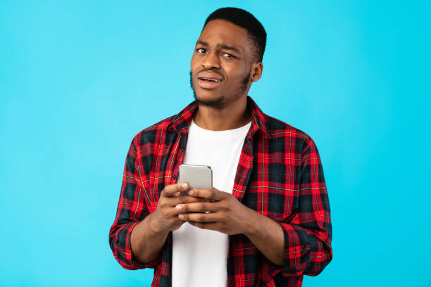 uomo nero incerto che tiene il cellulare in posa su sfondo blu - sadness men clothing disappointment foto e immagini stock