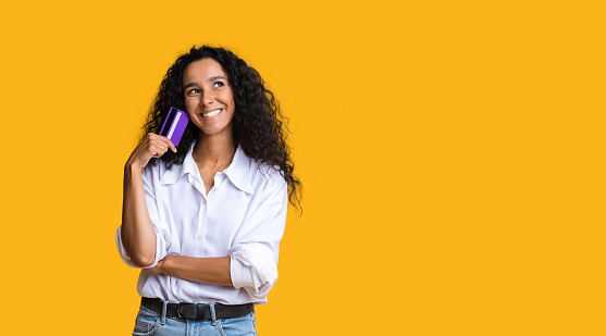 Shopping Time. Dreamy brunette woman holding credit card and looking away, posing over yellow studio background, panorama with copy space