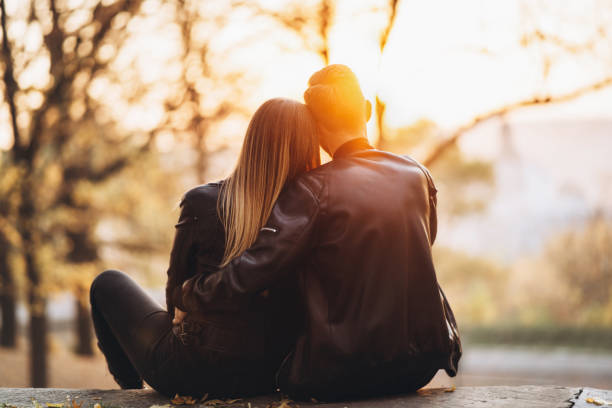 A romantic couple sitting on a bench hugging and watching the sunset in the park. Love story. Back view stock photo
