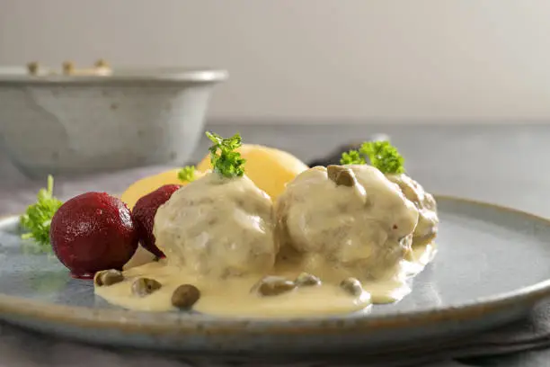 Boiled meatballs in white sauce with capers, in Germany called Koenigsberger Klopse, traditional dish with beetroot potatoes and parsley garnish on a gray plate, copy space, close-up with selected focus and narrow depth of field