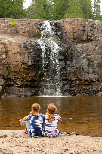 Two young girls (sisters) sitting side by side as they watch a waterfall at Gooseberry Falls State Park in Minnesota, USA.