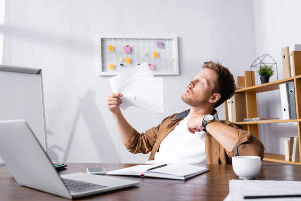 istock Selective focus of young businessman blowing with documents while suffering from heat at table in office 1271734134