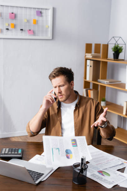 istock Selective focus of skeptical businessman talking on smartphone and throwing papers in office 1271734138
