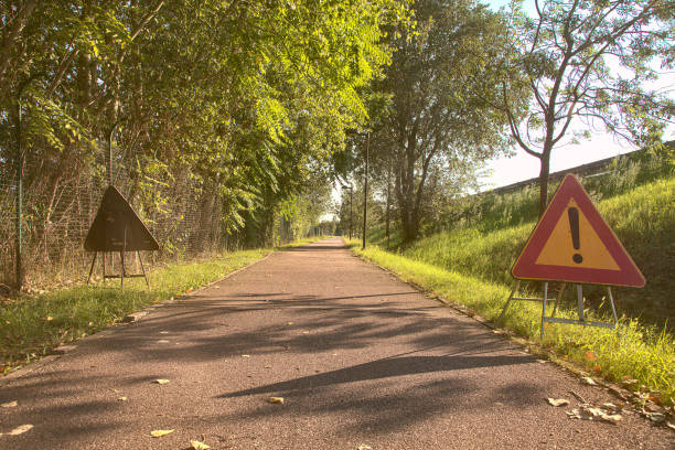 ciclovia no campo com árvores e luzes de rua e uma placa de perigo no chão em um dia claro no verão - single lane road road sunset rural scene - fotografias e filmes do acervo