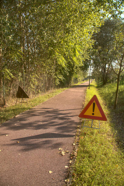 ciclovia no campo com árvores e luzes de rua e uma placa de perigo no chão em um dia claro no verão - single lane road road sunset rural scene - fotografias e filmes do acervo