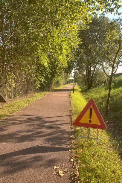 ciclovia no campo com árvores e luzes de rua e uma placa de perigo no chão em um dia claro no verão - single lane road road sunset rural scene - fotografias e filmes do acervo