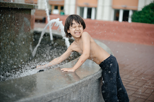 A little boy without a shirt is bathing in a fountain. Dark wet hair. The concept of happiness and positive. Carefree childhood and summer.