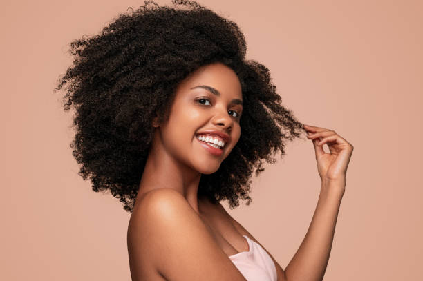 Happy black woman touching clean hair Side view of positive African American female model smiling for camera and touching clean curly hair after hygienic routine against brown background curly hair stock pictures, royalty-free photos & images