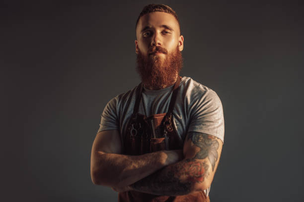 Confident modern barber looking at camera Contemporary bearded lumbersexual hairdresser crossing arms and looking at camera against gray background hipsters stock pictures, royalty-free photos & images