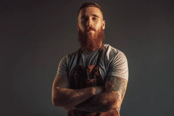 Contemporary bearded lumbersexual hairdresser crossing arms and looking at camera against gray background