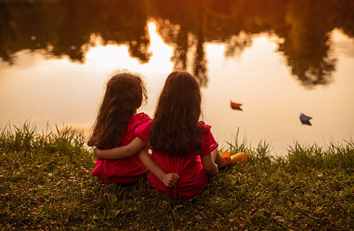 Back view of anonymous sisters hugging each other while sitting on grassy coast near calm lake water in evening in park