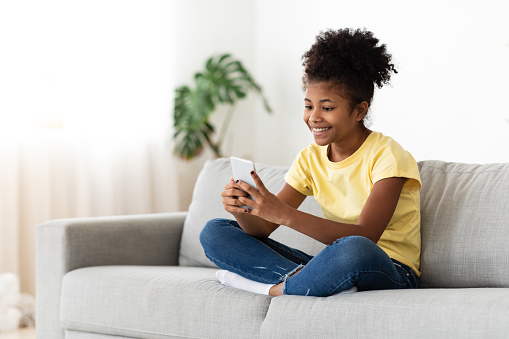 Mobile Games. Black Kid Girl Playing Online On Smartphone Sitting On Couch At Home. Empty Space