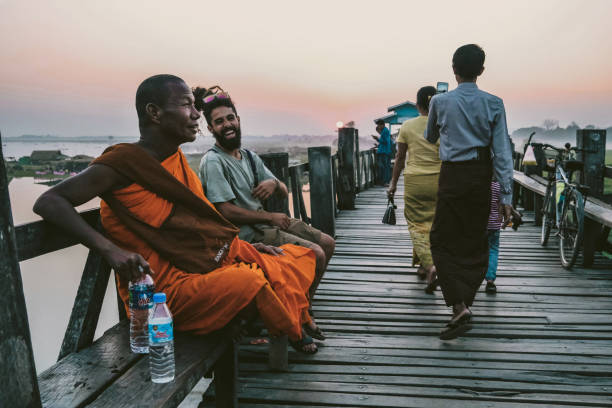 The genial Monk at the Mandalay Ubein Bridge. Access to the wooden bridge from popular destinations. People on the walk and tourists with dreadlocks smiling next to Monk.The orange Monk suit is the old buddsit Monk. He grins cross-legged on the bridge. Water bottle in hand. Lake in the back. Location: Ubein bridge / Mandalay u bein bridge stock pictures, royalty-free photos & images