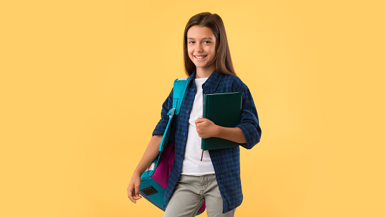 Smiling student girl wearing school backpack and holding exercise book, smiling at camera, pastel orange wall
