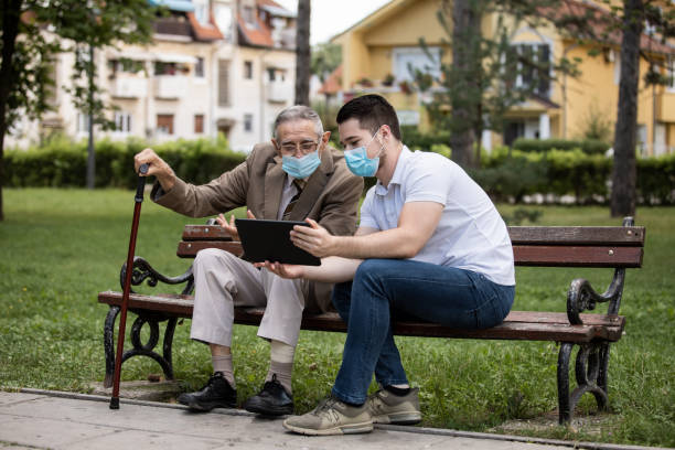 Two generations, a grandson and a grandfather, sit on a bench using a tablet and talking under masks