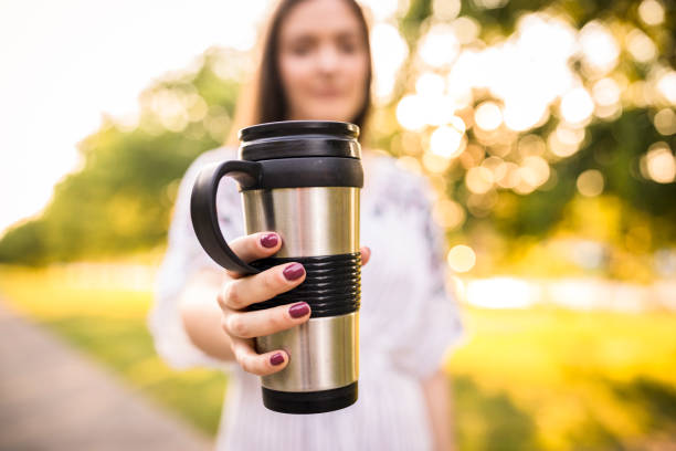 Close up shot of young woman holding her reusable coffee mug and showing it to camera while standing in a sunny public park.