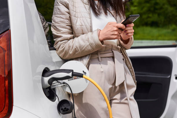jeune femme debout près de la voiture électrique avec le téléphone portable dans sa main et attendant la recharge de la batterie d’automobile. - electrical conduit photos et images de collection