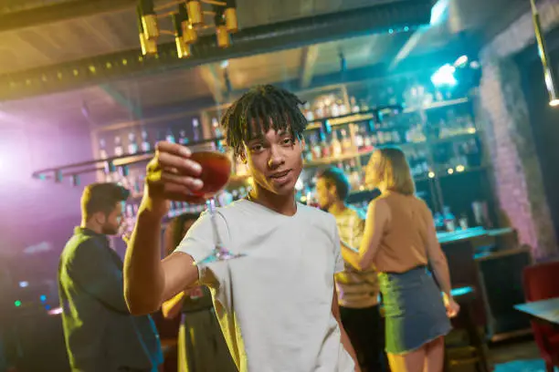 Photo of More Vibe, More Fun. Cheerful mixed race young man getting drunk, posing with a cocktail in his hand. Friends celebrating, chatting, having fun in the background