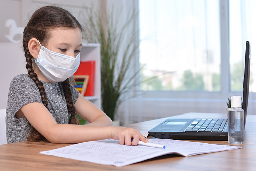 A schoolgirl in a medical mask is doing her homework on a laptop at home or studying in class at a lesson at school.Student safety after covid-19 pandemic. Back to school.Distance learning.