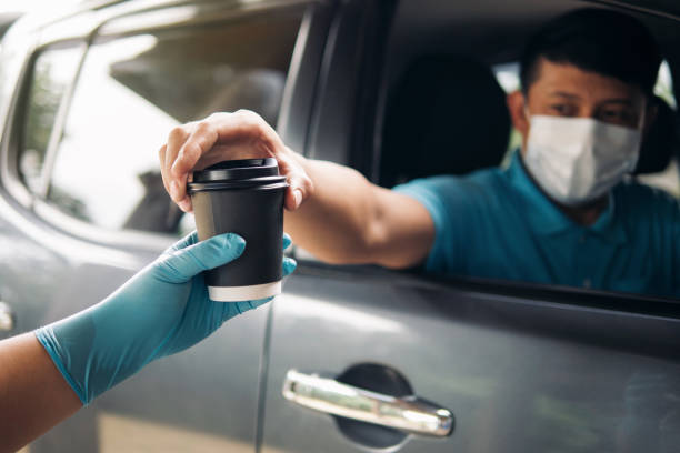 Drive thru food and drink service during Coronavirus COVID-19 pandemic. Staff hand in protective glove serving a coffee cup to customer a car driver man in protective face mask at drive thru service. conduzca a través del servicio de comida y bebida durante la pandemia de coronavirus covid-19. el personal de la mano en guante de protección que sirve una taza de café al cliente un conductor de coche con máscara protectora en la unidad a través del