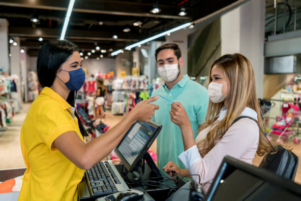 couple shopping at a clothing store and using facemasks during the pandemic - clothing store paying cashier credit card imagens e fotografias de stock
