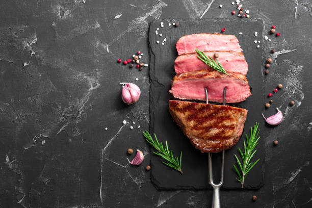 istock Medium rare Ribeye steak on a fork with spices and herbs on a black stone background. BBQ beef fillet cut into pieces. Top view, flat lay, copy space. 1271776660