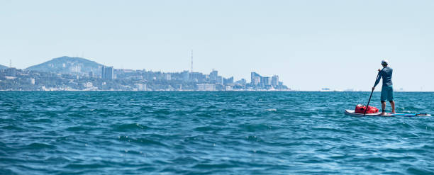 Active man rowing on SUP touring with waterproof bag at Sochi city coastline background