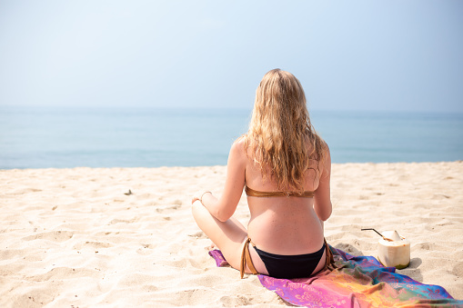 Caucasian woman in bikini sits on the sand on the beach in the lotus position. Nearby there is a coconut for drinking. Travel and tourism. Yoga classes.
