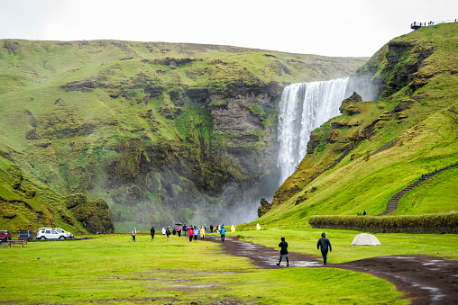 Skogafoss, Iceland waterfall on cliff with green grass field and many tourists people walking with umbrellas and ponchos on trail road path to water fall