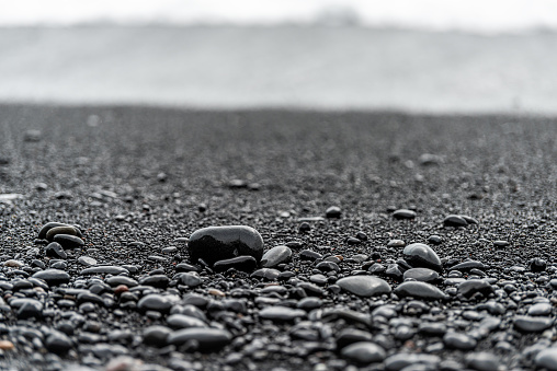 Closeup foreground of large black volcanic stones or pebbles on sand beach in Reynisfjara, Iceland with water waves crashing on shore to shiny wet rocks