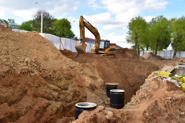 Photo of Excavator on earthwork during the laying of pipes of the heating system to a new residential building at the construction site. Laying concrete sewer wells, rings and manholes in ground.