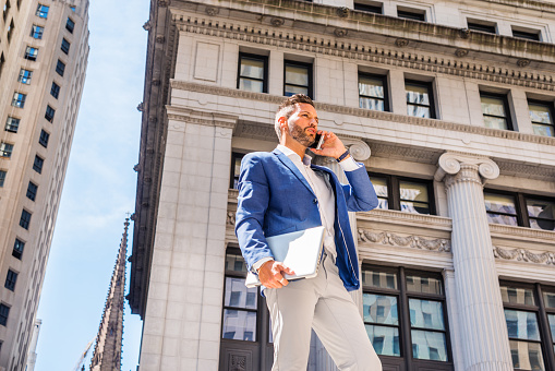 Young European Businessman with beard traveling, working in New York City, wearing blue blazer, gray pants, holding laptop computer, standing outside old office building, calling on cell phone.
